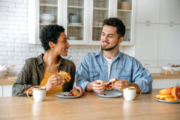 Fototapeta na wymiar Positive couple in love of different nationalities, happy spouses, sit at home in the kitchen, drinking morning coffee with croissants with jam, look at each other with love, smile