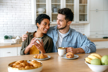 Happy young family couple, caucasian man and african american woman, having breakfast at cozy home kitchen with croissants and coffee, look at each other, smile. Sunday morning together