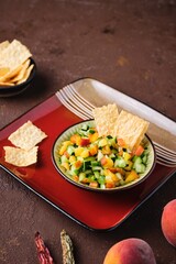 Salsa from fresh cucumber and ripe peach in a ceramic plate on a brown concrete background. Served with wheat chips. Mexican cuisine.
