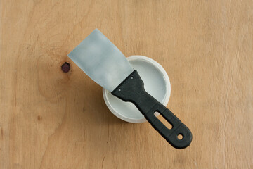 Construction putty knife and can of white putty on plywood surface. Still life. Putty application kit. View from above.
