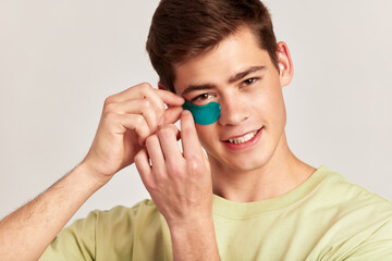 A millennial guy with brown hair practicing skincare with under eye hydrogel patches in a studio. Young smiling caucasian man applying cosmetic patches on his face. Men's self-care.