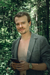 Black and white portrait of Self assured smiling young handsome man with naked chest and open grey jacket. Macho with dark hair, sideburns and beard with phone in park, looking at camera. 