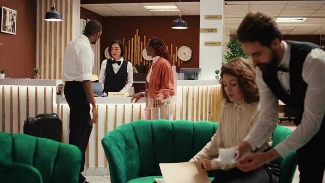 Busy hotel manager receiving cup of coffee from employee while checking paperwork of guests waiting to be checked in. Receptionist offers room access to couple after booking process is finalized