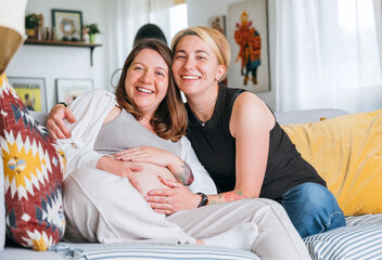 Happy smiling women couple waiting for baby. Same-sex pregnant marriage couple on home living room...