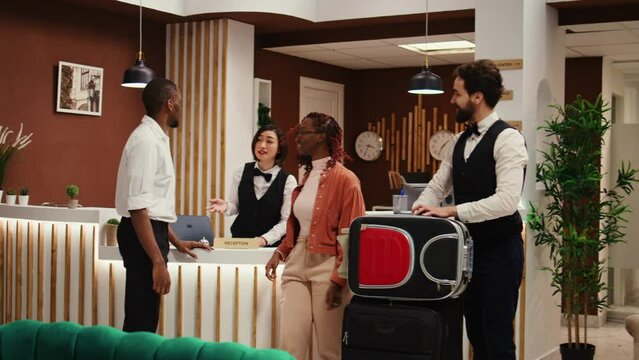 Guests checking out from resort, handing over room access card key while friendly bellboy helps them with their baggage. Happy relaxed tourists leaving hotel after nice stay