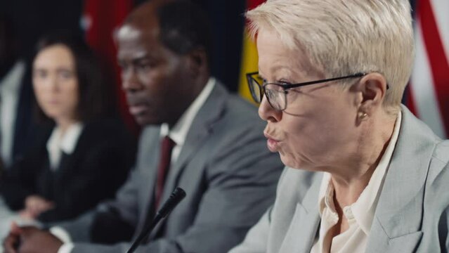 Close-up selective focus shot of furious middle-aged blonde Caucasian female politician talking at international crisis summit, diverse heads of state listening, various flags in blurred background