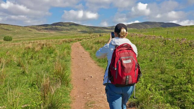 Behind a backpacker taking a picture with her phone while walking at the Scottish Rolling West Highland hills on a trail, Scotland