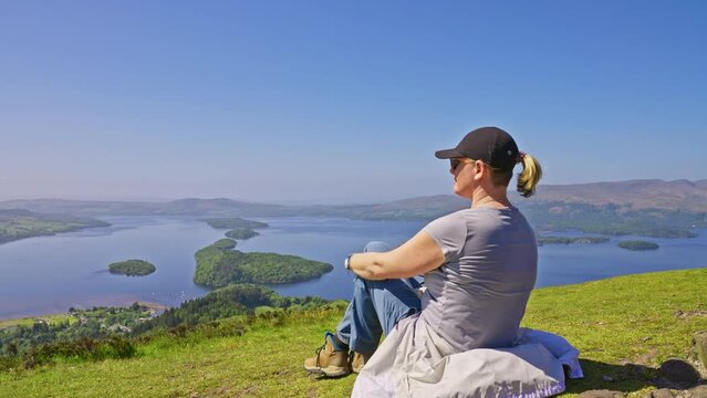 Parallax tracking a woman sitting on a grassy hillside with rolling hills and view of the lack in the distance, Conic Hill, Scotland