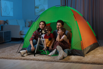 Happy family using telescope to look at stars while sitting in camping tent indoors