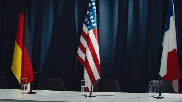 Pan shot of empty conference room prepared for international meeting, with flags of Canada, France, USA, Germany, tables, chairs, microphones, glasses of water, and dark blue drapes in background