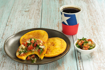 Sopaipillas with bite of pumpkin or squash and pebre, on a round plate with a poly paper cup with Chilean flag, on a wooden table and copy space. concept of Chilean national holidays