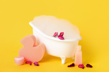 Small bathtub with foam, flower petals and bath supplies on yellow background