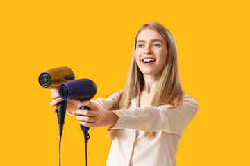 Young blonde woman in pajamas with hair dryers on yellow background