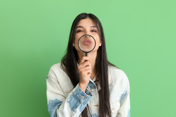 Beautiful young woman with magnifier blowing kiss on green background