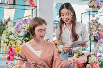 Two young beautiful female florist partners discuss bloom bouquet arrangement design in tablet, online purchase order work in colorful flower shop store with fresh flora, SME business entrepreneur.