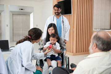 Happy young female pediatric doctor in uniform teasing little boy in wheelchair for medical exam at...