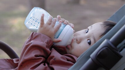 Asian baby drinks formula from a bottle while lying on the stroller in the park