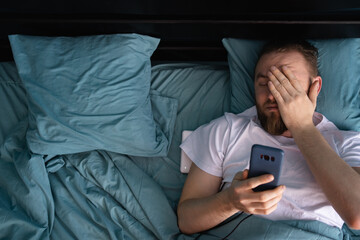 Confused man looking on mobile phone lying in bed at home in the morning. Oversleeping concept.