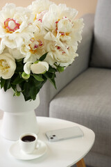 Vase of white peonies with cup of coffee and mobile phone on table near couch