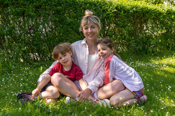 Naklejka na ściany i meble Modern mom sits on the grass and hugs cute little children,son and daughter. Cheerful family, joyful children. outdoor time with kids