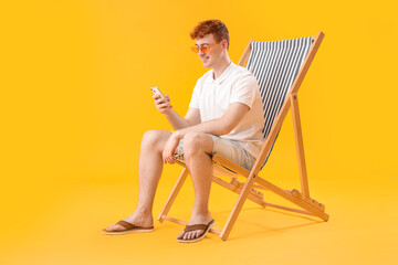 Young man using mobile phone in deck chair on yellow background