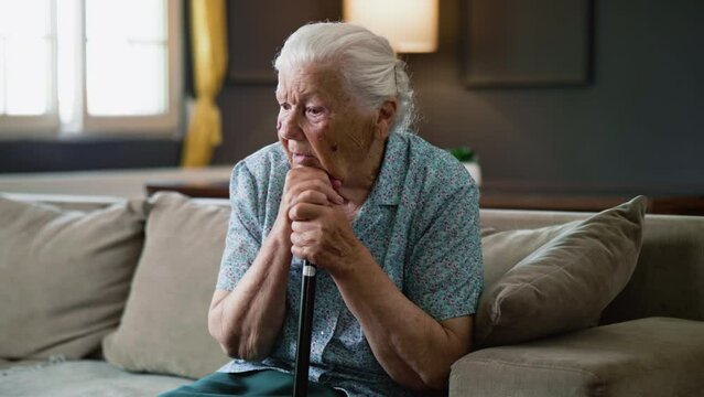 Upset senior retired 80s woman grandmother holding cane stick in hands sit alone depressed on sofa at home
