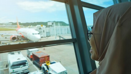 Asian woman wearing hijab smiling looking at airplane while waiting for departure at the airport. Muslim traveler and vacation concept