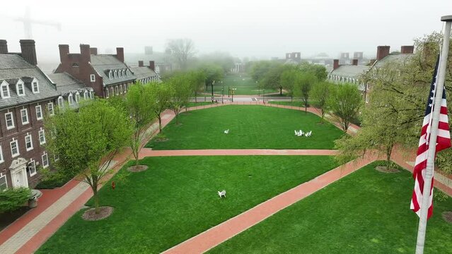 Aerial drone videography of American flag flying above college campus. Historic brick academic buildings line green quad with seating for students. Foggy damp morning with university theme.
