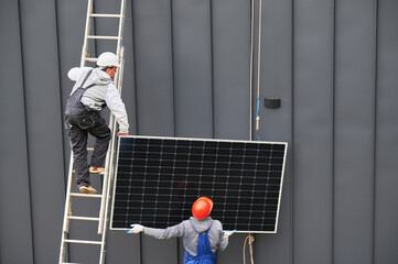 Men mounters installing solar panel system on roof of house. Workers in helmets lifting up photovoltaic solar module with help of ropes outdoors. Concept of alternative and renewable energy.