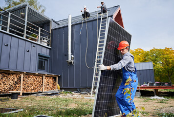 Builders building solar panel system on roof of house. Man worker in helmet carrying photovoltaic...