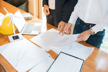 Close-up of hands of two Asian male architects pointing at blueprints on a desk. about construction industry design Includes a white windmill model, a laptop, a pen, and a helmet. Placed in the office