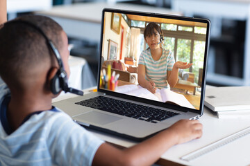 Fototapeta na wymiar Diverse schoolboy with headphones and schoolgirl at desk having laptop video call