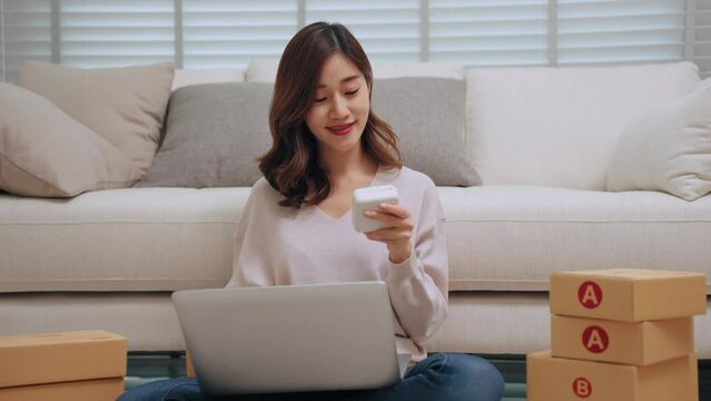 Young asian woman selling online receiving order via laptop and using label printer to printing customer address attach to the parcel box. Business Online and delivery shop concept