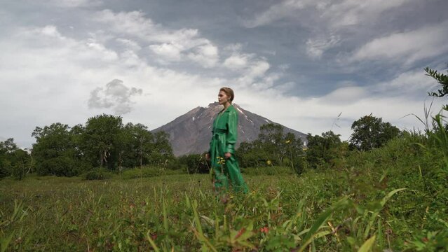 Attractive blonde female model dressed in green leather coat walking in forest against backdrop of volcano on summer day. Handheld, wide low angle shot, scenic view. Side view. Part of video series