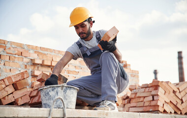 Bucket on the ground, holding a brick. Handsome Indian man is on the construction site