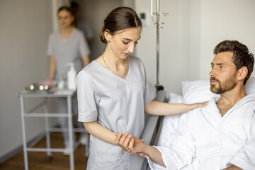 Obraz premium Young nurse takes care of adult man sitting on bed of comfortable medical ward. Medical support and assistance during the rehabilitation in clinic