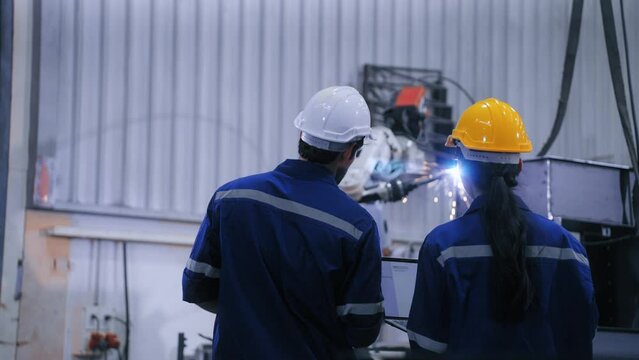 Two professional engineers control manage robot arm welding automation in heavy industry. Skillful man and woman working by welding robot in steel factory. Manufacturing automatic system concept