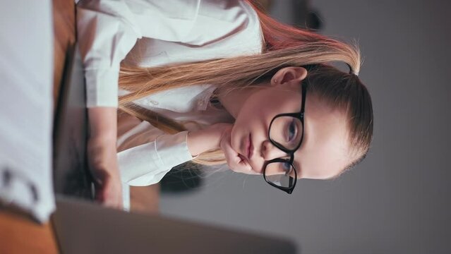Close up of delightful schoolgirl in black trendy glasses holding hand under chin and looking at laptop screen indoors. Adorable child turning eyes on camera and making gentle smile. Vertical video.