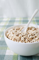 Raw oatmeal in a white bowl on a green checkered tablecloth with a teaspoon. Concept of healthy food. Vegetarian and vegan. Vertical orientation. Selective focus.