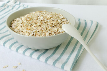 Raw oatmeal in a gray bowl on a white tablecloth with a kitchen napkin and a teaspoon. Concept of healthy eating. Vegetarian and vegan food. Horizontal orientation. Selective focus.