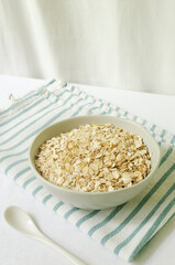 Raw oatmeal in a gray bowl on a white tablecloth with a kitchen napkin and a teaspoon. Concept of healthy eating. Vegetarian and vegan food. Vertical orientation. Selective focus.
