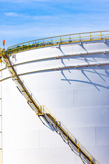 White oil storage fuel tanks at depot station with access ladder against a blue sky. Vertical fuel tank staircase.Oil and gas industry.curve line spiral staircase on storage fuel tank.Oil industry,