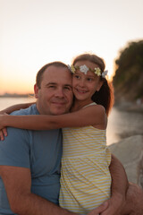 happy father with his cute daughter hugging on the seashore. girl in a yellow dress with a wreath on her head radiating happiness and love.