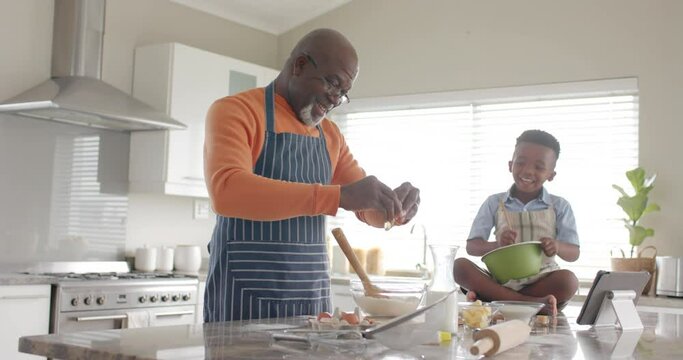 Happy african american grandfather and grandson baking, using tablet in kitchen, slow motion