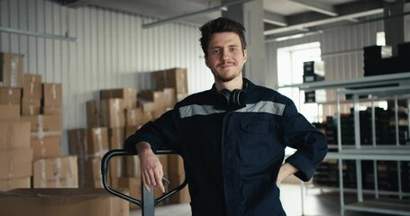 A man in a blue uniform smiles and looks into the camera, leaning on the handle of a special machine against the backdrop of a warehouse at a factory