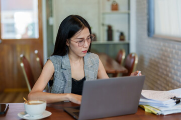 Asian woman sitting working in cafe, there is laptop computer in foreground large pile documents, was busy with pile paperwork, looking for document that needed be solved, have serious expression.