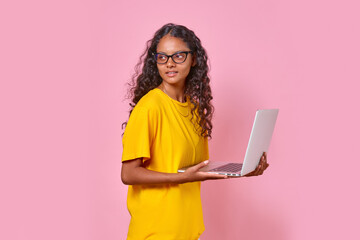 Young beautiful attractive Indian woman student holding open laptop and preparing for high school exams via internet or getting online education dressed in casual clothes stands on pink background.