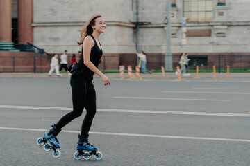 Fototapeta premium Athletic woman on roller skates moves actively poses on asphalt at street dressed in active wear has cheerful expression. Young fitness model skating in urban place. Sporty lifestyle rollerblading