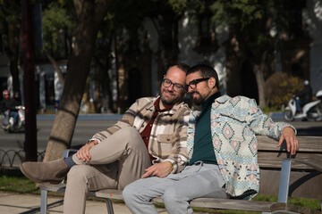 Fototapeta premium Real marriage of gay couple, holding hands, heads together and eyes closed, sitting on a wooden bench, complicit and happy. Concept lgtb, lgtbiq+, couples, in love, complicity.