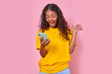 Young overjoyed ethnic Indian woman teenager holds mobile phone with school app and screams happily seeing message about highest mark for exam stands in pink studio. Training, education, knowledge